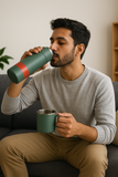 Man sitting on a couch drinking from a green thermos.
